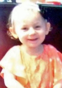 Photo of Kendall Chick, a toddler girl wearing an orange dress. Her pale skin is washed out by sunlight, and she is wearing a hair ribbon with a black flower in her wispy blonde hair. She is smiling.