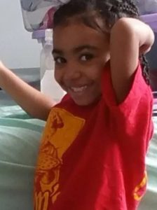 Photo of a young African-American girl wearing an oversized red T-shirt. Her hair is done in braids; she has an arm behind her head, and she is smiling.