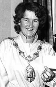 Black and white photo of Janette Dunbavand as a younger woman. She has short curly dark hair and pale skin; she is wearing a white robe and a mayor's chain of office.