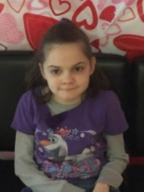 Photo of Ashlyn Ellis, a young girl with long brown hair, looking at the camera, biting her lower lip. She is wearing a purple sweater.