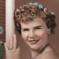 Colorized black-and-white photo of a young woman with curly hair, fair skin, and blue flowers in her hair, smiling for the camera. She has an engagement ring on her finger.