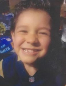 Photo of a small boy with tan skin and curly brown hair, grinning at the camera with his eyes nearly shut.