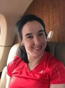 Photo of Rebecca Fogel, a young woman sitting in an airplane seat, wearing a red T-shirt and smiling for the camera. Her messy dark-brown hair is drawn back in a half-ponytail.