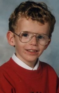 Photo of a small boy wearing large wire-framed glasses and a school uniform with a white collar under a red sweatshirt. He has pale skin and curly blond hair. Mild bruises are visible on his lower lip. He is smiling for the camera.