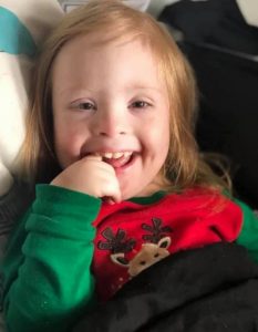 Photo of Kylee Willis, a toddler girl with Down syndrome. She is wearing a Christmas sweater with a reindeer on it. She is grinning and has her forefinger in her mouth. Her hair is light blonde and wispy.