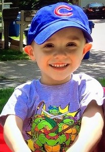 Photo of a small boy in a big blue baseball cap and Teenage Mutant Ninja Turtles T-shirt. He has pale skin and his ears stick out, and he is smiling, showing he still has all his baby teeth.