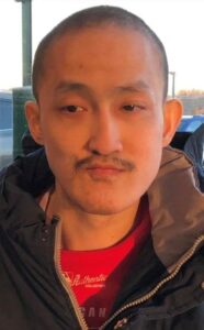 Photo of an Asian man with light skin, black hair in a buzz cut, and a thin black mustache, wearing a brown jacket over a red shirt.