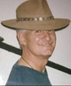 Photo of a fair-skinned man, smiling for the camera, wearing a wide-brimmed tan hat and teal shirt.