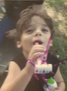 Lukas, a child with frizzy brown hair and olive-toned skin, is about to blow into a pink birthday blower tied with pink ribbons. He is wearing a black shirt.