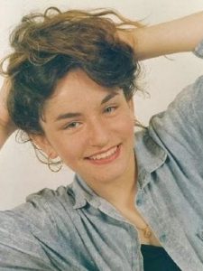 Tilted photo of a young woman with messy brown hair, wearing a denim jacket and smiling at the camera.