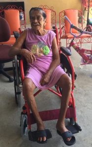 Thin elderly woman with dark hair and light-brown skin, dressed in a pink shirt and shorts, smiling for the camera, sitting in a red wheelchair.