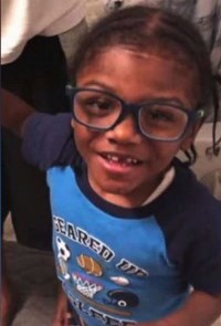 A small boy with thick glasses, front teeth missing, hair in cornrows, wearing a soccer shirt. He has dark-brown skin and black hair.