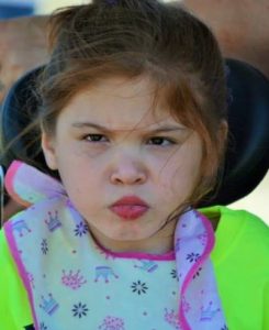 Photo of a young girl sitting in a wheelchair, lips pursed, looking annoyed. She has fair skin and her brown hair is messy.