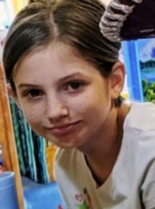 Photo of a girl with light, freckled skin, brown hair pulled back, smiling for the camera.