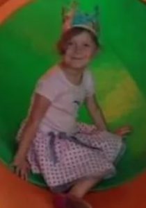 Photo of a girl in a polka-dot skirt and gold plastic crown, sitting in a green play tunnel; she has short strawberry-blond hair and fair skin.