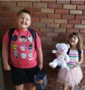 Photo of Conner and Brinley Snyder. He is a heavyset boy wearing a T-shirt, shorts, and backpack; she is a girl wearing a frilly pink skirt and holding a white teddy bear.