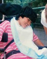 Photo of a teenage girl with light skin and short dark hair sitting in a wheelchair, looking off-frame and smiling at something at knee level. She is wearing a red and white striped shirt, and a disposable cloth is draped around her neck.