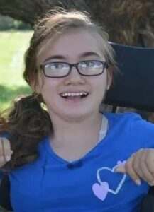 Photo of a teenage girl with a dark-blonde ponytail and glasses, sitting in a wheelchair.