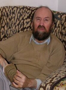 Photo of a middle-aged man in a beige cardigan, sitting in a living chair and reading a book. He has light skin, balding black hair and a beard.