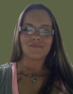 Photo of a young woman with long, straight brown hair and fair skin, wearing glasses. She is wearing a gray sweater over a pink shirt, and a chain around her neck holds a silver ring. She has a script tattoo along her collarbone.