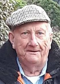 Photo of a fair-skinned elderly man with a tweed cap and wool jacket, photographed outside.