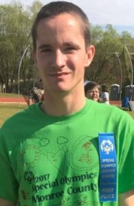Photo of a young man in a green Special Olympics T-shirt; he has a buzz cut and is squinting into the sun.