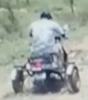 Blurred photo, taken from behind, of a person in a white shirt riding a three-wheeled scooter down a dirt road.