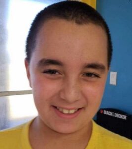 Photo of a boy smiling for the camera. He has short-cropped brown hair and is wearing a yellow shirt.