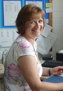 Photo of a middle-aged woman sitting at a desk; she has brown, curly hair and fair skin.