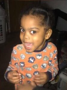 Photo of a small boy with brown skin and black hair, wearing an orange print pajama top.