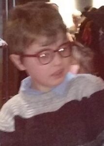 Blurry photo of a young boy wearing a sweater and thick glasses. His dirty-blond hair is parted on the side and his expression is neutral.