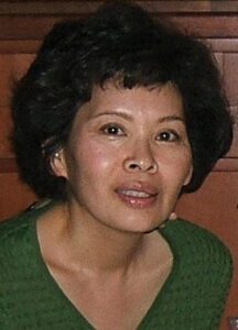 Middle-aged Asian woman, her short black hair permed, wearing a green shirt.
