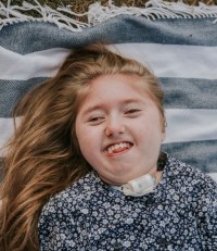 Photo of a girl lying on a striped blanket. Her skin is fair and her long hair is strawberry-blonde. She is smiling broadly and wearing a black-and-white flowered shirt and a trach collar.