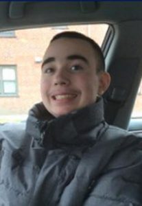 David Almond, a teenage boy, sits in a car and smiles for the camera. He is wearing a winter scarf and heavy gray parka. His hair is dark and very short, and his skin is pale brown.