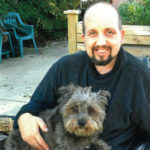 Photo of a middle-aged man, balding, with fair skin and a neat goatee, holding a small gray shaggy terrier on his lap.