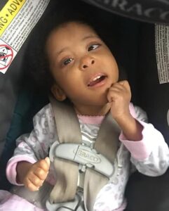 Photo of a toddler girl in a car seat. She is wearing a pink onesie and her hands are balled into fists. Her skin is tan and her eyes are dark brown.