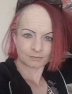 Photo of a woman with pale skin, thin eyebrows, and a punk haircut that is dyed pink, shaved down on the left side, and straight and chin-length on the right.