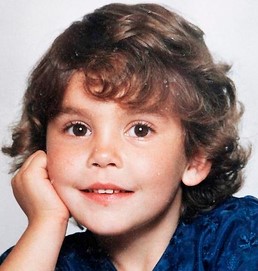 Phot of a small girl with fair skin, brown eyes, and curly brown hair. She is leaning her head against her hand and smiling for the camera.