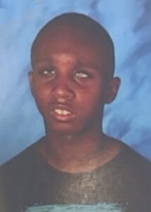 School photo of a boy with dark skin and very short hair, wearing a black T-shirt.