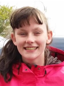 Photo of a teenage girl with wavy brown hair cut into bangs, gray eyes, and pale skin, smiling for the camera.