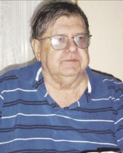 Photo of an older man with pale skin and gray hair, wearing glasses and a blue striped polo shirt.