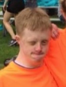 Photo of a young man with strawberry-blond hair and pale skin, wearing a fluorescent orange T-shirt and squinting in the sun.