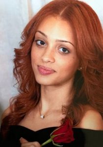 Photo of a woman with red hair, light-brown skin, and light make-up, wearing a black dress and holding a rose.
