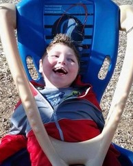 Photo of a smiling boy lying in an adapted swing. He has pale skin and curly brown hair; his face is turned up to the sun.