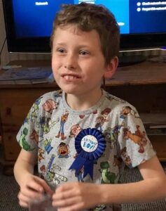 Photo of a young, sandy-haired boy wearing a blue "Birthday Boy" ribbon. His hands are blurred from waving them. 