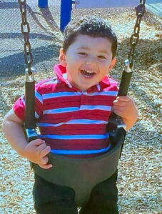 Photo of a small boy sitting in a swing and smiling broadly at the camera. He has black hair and tan skin and is wearing a striped red and blue shirt.