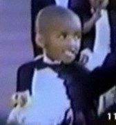 Blurry photo of a small boy in a suit; he has brown skin and a shaved head.