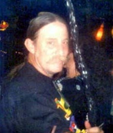 Photo of a middle-aged man with fair skin, his dark hair in a ponytail; he is wearing a black T-shirt.