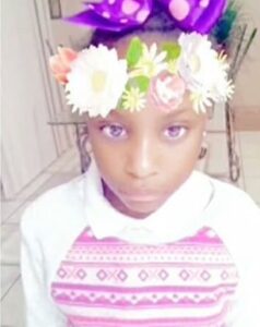 Photo of a young girl with brown skin, a pointed chin, and a solemn expression. A filter has been applied, giving her lavender eyes and flowers in her hair.