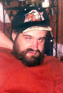 Photo of a man in a baseball cap printed with an eagle. He has dark hair, light skin, and a full beard. His shirt is red; he has his hands behind his head and is leaning back.